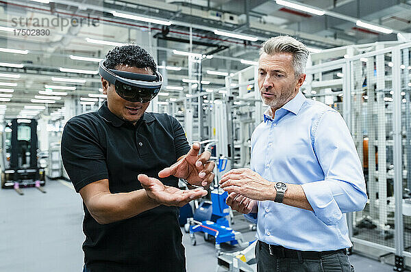 Geschäftsmann blickt auf Ingenieur mit Smart-Brille  der in Fabrik gestikuliert