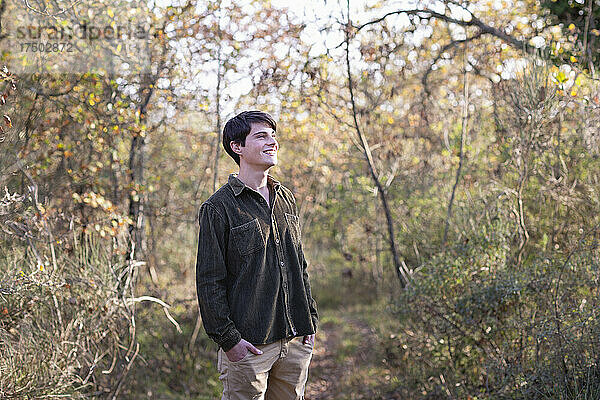 Lächelnder junger Mann mit den Händen in den Taschen  der in den Wald blickt