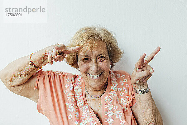 Glückliche ältere Frau gestikuliert Friedenszeichen vor der Wand