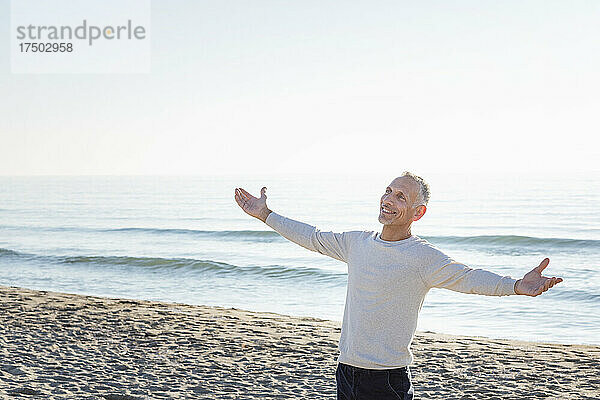 Lächelnder Mann mit ausgestreckten Armen steht am Strand