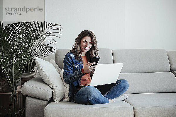 Lächelnde Frau mit Laptop und Smartphone auf dem Sofa