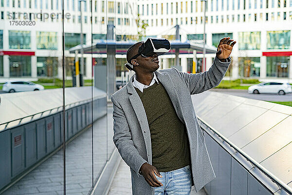 Geschäftsmann gestikuliert mit Virtual-Reality-Headset in der Nähe einer Glaswand in der Stadt