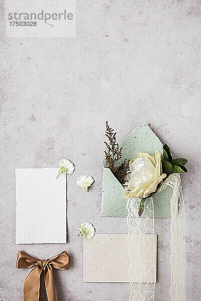 Studioaufnahme eines Umschlags  einer Einladungskarte  einer gebundenen Schleife und flach gelegter Blumen vor hellrosa Hintergrund