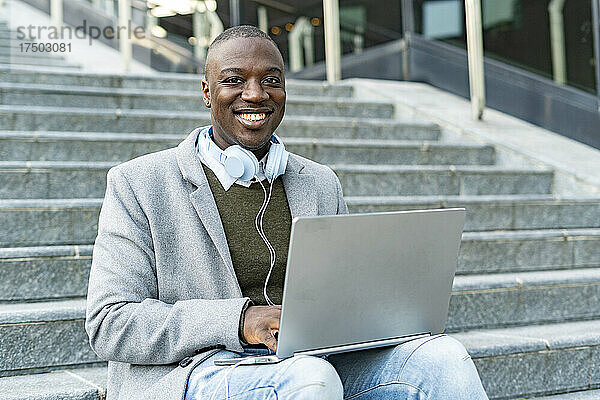 Lächelnder Geschäftsmann mit Kopfhörern und Laptop sitzt auf Stufen