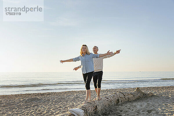 Paar mit ausgestreckten Armen läuft auf Baumstamm am Strand