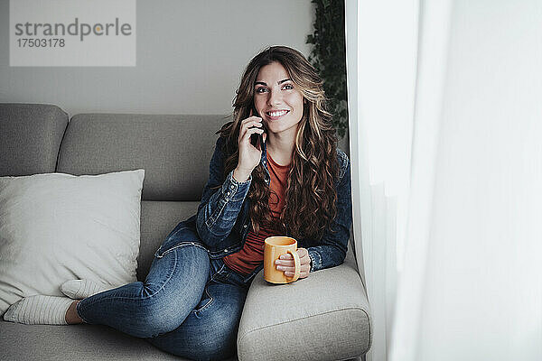 Lächelnde Frau mit Kaffeetasse  die zu Hause auf dem Smartphone spricht