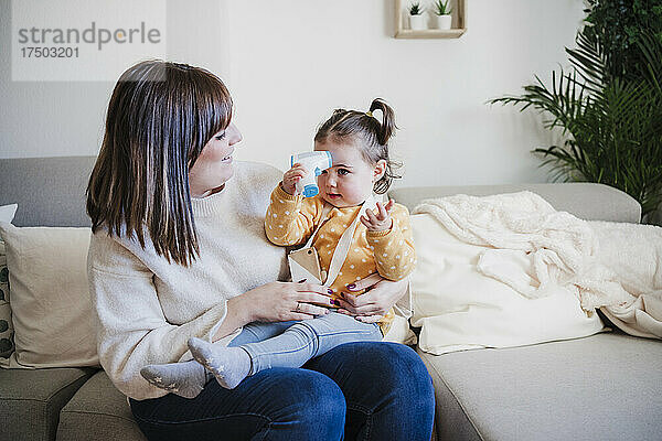 Lächelnde Mutter blickt auf süßes Baby  das ein Infrarot-Thermometer auf dem Sofa hält