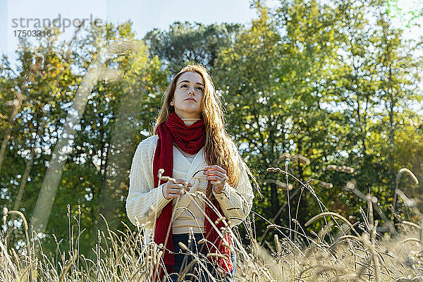 Nachdenkliche Frau mit rotem Schal steht an einem sonnigen Tag inmitten von Gras