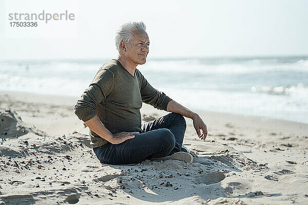 Lächelnder älterer Mann sitzt mit gekreuzten Beinen im Sand am Strand