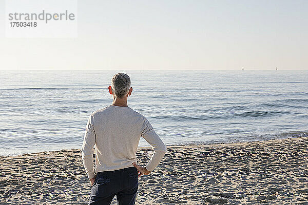 Mann mit der Hand auf der Hüfte und Blick auf das Meer  der am Strand steht