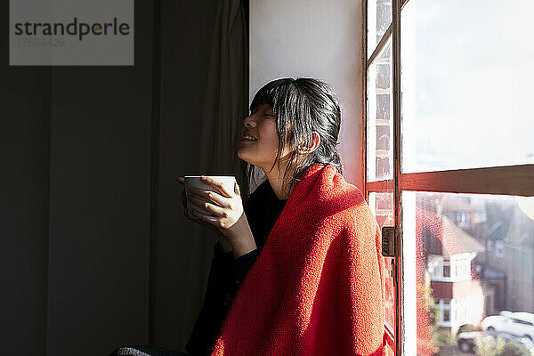 Frau mit Decke hält Kaffeetasse am Fenster zu Hause