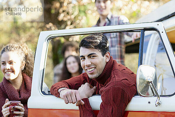 Lächelnder junger Mann lehnt mit Freunden an der Tür eines Lieferwagens und genießt ein Picknick im Herbstwald