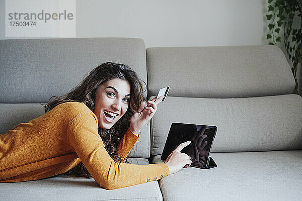 Aufgeregte Frau benutzt Tablet-PC und hält Kreditkarte zu Hause auf dem Sofa