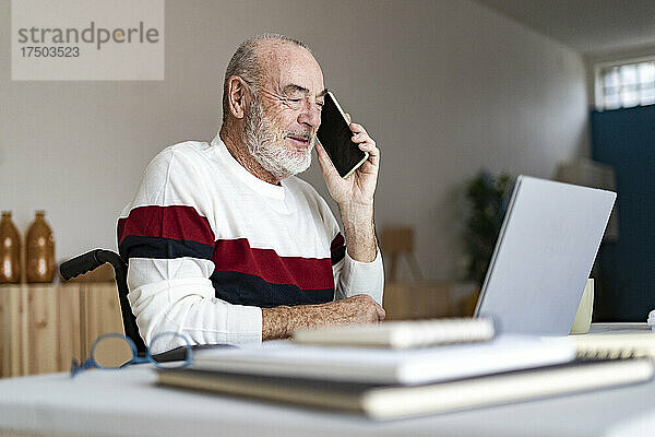 Älterer behinderter Geschäftsmann mit Laptop  der zu Hause mit dem Handy telefoniert