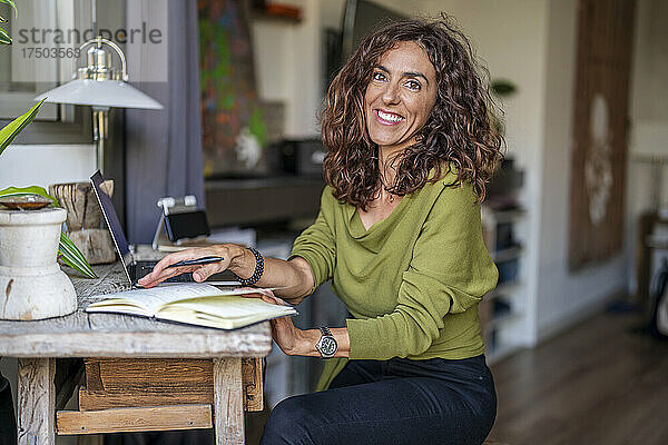 Lächelnde Frau mit Tagebuch und Laptop am Tisch