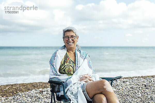 Lächelnde ältere Frau  in eine Decke gehüllt  sitzt am Strand