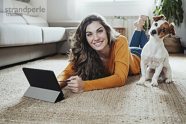 Frau mit Tablet-PC liegt mit Hund zu Hause auf dem Boden