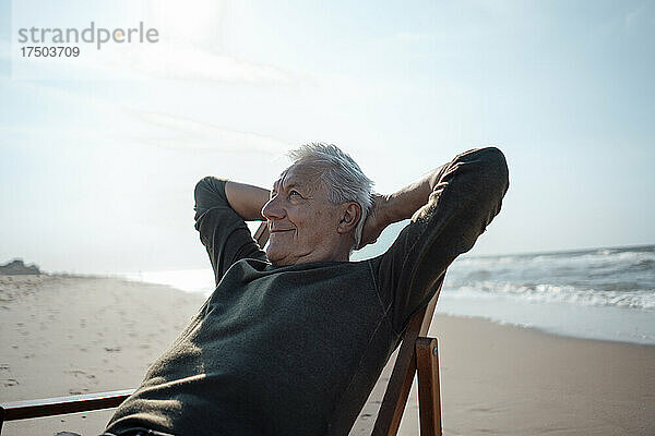 Lächelnder älterer Mann mit den Händen hinter dem Kopf entspannt am Strand