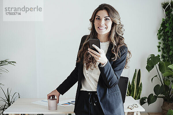 Lächelnde Geschäftsfrau mit Smartphone steht neben Schreibtisch im Büro