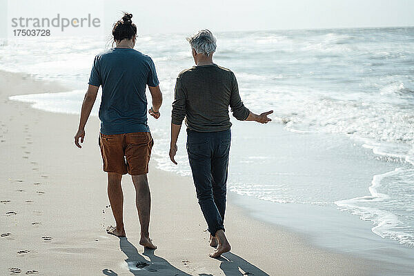 Älterer Mann gestikuliert mit seinem Sohn  der gemeinsam am Strand spazieren geht