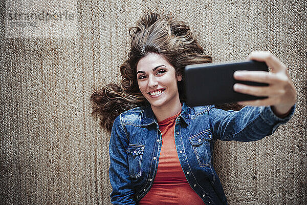 Lächelnde junge Frau mit Mobiltelefon macht ein Selfie auf dem Teppich