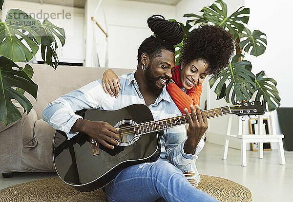 Frau mit lockigem Haar umarmt ihren Freund  der zu Hause Gitarre spielt