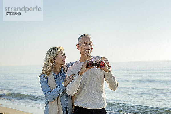 Lächelnde Frau mit Arm um Ehemann und Fernglas am Strand