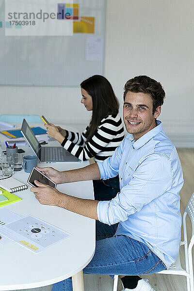 Lächelnder Geschäftsmann mit Tablet-PC sitzt mit Kollegen im Büro und nutzt Smartphone