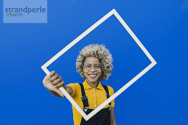 Frau mit Afro-Frisur hält Rahmen vor blauer Wand