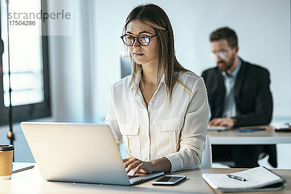 Junge Geschäftsfrau mit Brille arbeitet am Laptop mit Kollegin im Hintergrund im Büro