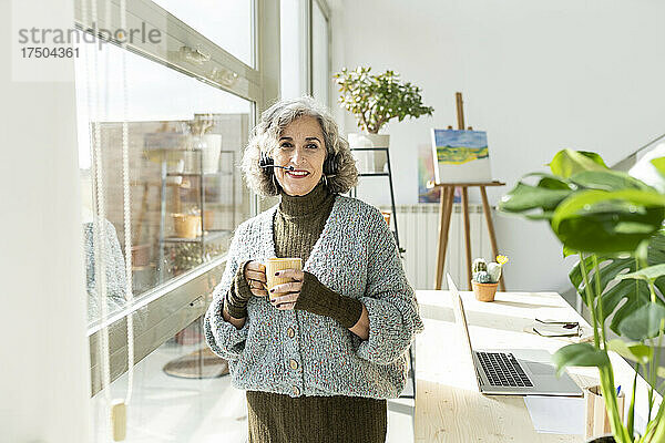 Lächelnde ältere Geschäftsfrau hält einen Becher in der Hand und steht im Heimbüro am Tisch