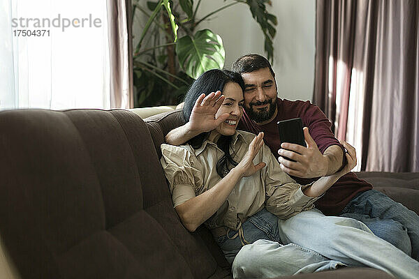 Paar winkt per Hand zu Videoanruf über Mobiltelefon auf dem Sofa