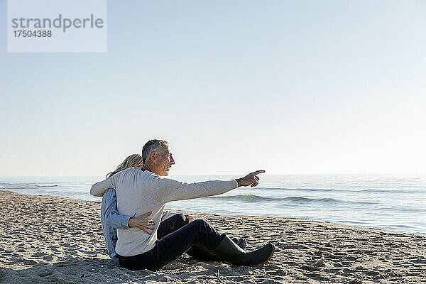Mann zeigt aufs Meer und sitzt mit Arm um Frau am Strand