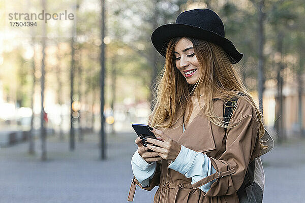 Lächelnde Frau im Trenchcoat benutzt Mobiltelefon in der Stadt