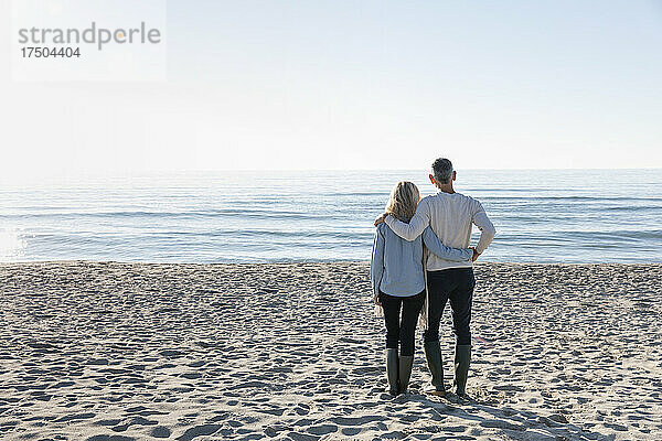 Mann mit Arm um Frau  die auf Sand steht und auf das Meer blickt