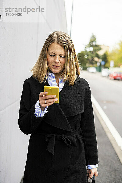 Lächelnde Frau  die über ihr Smartphone SMS schreibt und an der Wand vorbeigeht