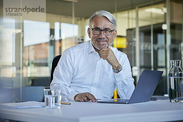 Lächelnder Geschäftsmann mit Laptop  der am Tisch im Büro sitzt