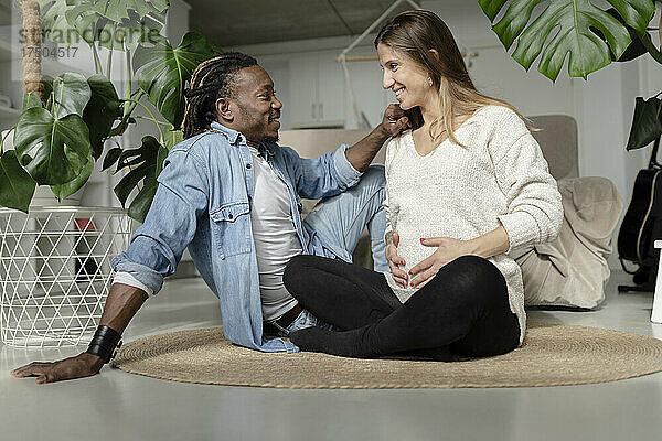 Lächelnder Mann blickt schwangere Frau an  die im Wohnzimmer ihren Bauch auf dem Teppich berührt