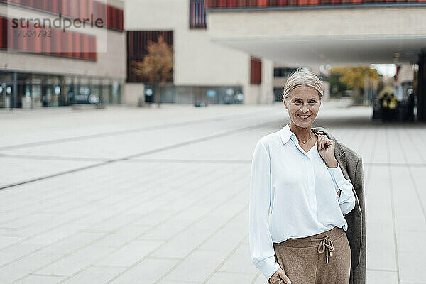 Lächelnde Geschäftsfrau mit der Hand in der Tasche steht auf dem Fußweg