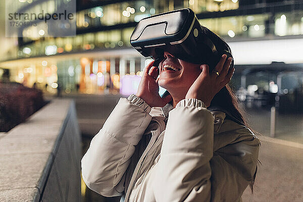 Glückliche junge Frau  die nachts ein Virtual-Reality-Headset an der Wand nutzt