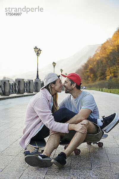 Romantisches Paar reibt sich die Nasen und sitzt auf dem Skateboard am Fußweg