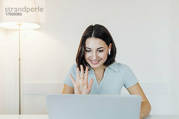 Lächelnde junge Geschäftsfrau winkt bei Videoanruf über Laptop im Heimbüro