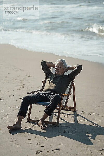 Älterer Mann mit den Händen hinter dem Kopf auf einem Stuhl am Strand