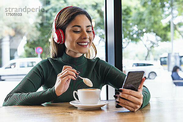 Lächelnde Frau mit kabellosen Kopfhörern  die im Café ihr Mobiltelefon benutzt