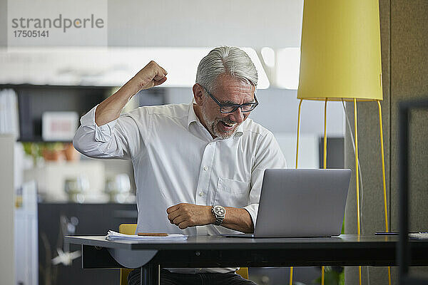 Geschäftsmann jubelt  während er am Arbeitsplatz per Videoanruf über Laptop spricht