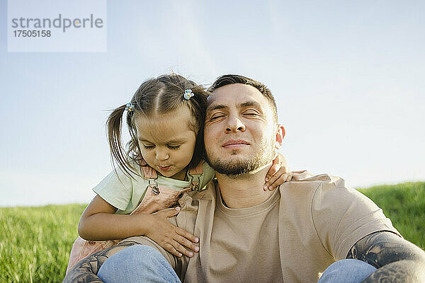 Mädchen umarmt Vater  der mit geschlossenen Augen auf der Wiese sitzt