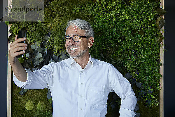 Lächelnder reifer Mann macht Selfie mit Smartphone vor Pflanzen im Büro