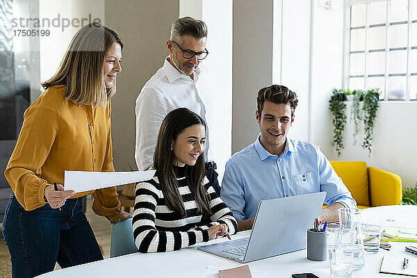 Geschäftsfrau mit Dokument schaut auf Laptop  während Kollegen im Büro diskutieren
