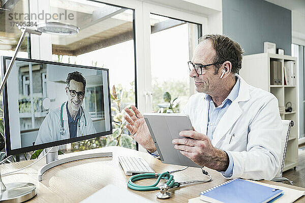 Arzt mit digitalem Tablet gestikuliert bei Videoanruf über Computer im Krankenhaus