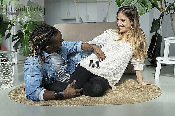 Lächelnde schwangere Frau neben einem Mann  der einen Ultraschallscan in der Hand hält und auf dem Teppich im Wohnzimmer sitzt